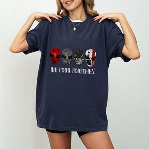 May include: Navy blue t-shirt featuring four stylized skull masks in red, gray, black, and white. The text "THE FOUR HORSEMEN" is printed below the masks in white. The shirt is a casual, oversized fit.