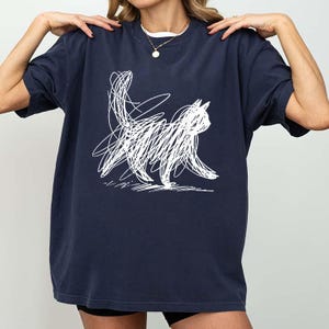May include: Navy blue t-shirt featuring a white scribbled cat design. The cat is depicted in a minimalist, abstract style, with a playful, artistic aesthetic. The shirt is a casual, comfortable garment.