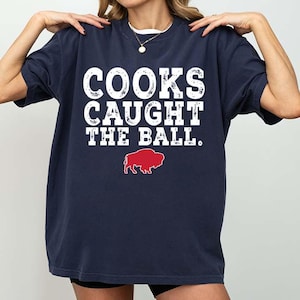Könnte beinhalten: Marineblaues T-Shirt mit dem weißen Text "COOKS CAUGHT THE BALL". Unter dem Text befindet sich eine rote Bison-Grafik. Das Shirt hat einen lässigen Stil.