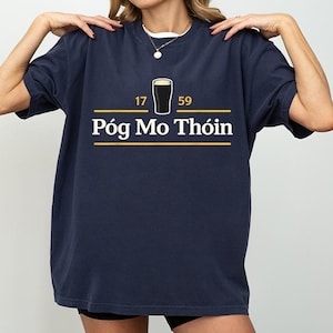 May include: Navy blue t-shirt with a graphic of a pint of stout and the text "Póg Mo Thóin" in white and yellow. The numbers "17" and "59" are on either side of the pint. The shirt is a relaxed fit.