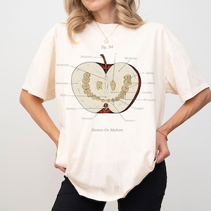 May include: Cream-colored t-shirt featuring a detailed anatomical illustration of an apple, labeled with scientific terms. The apple's interior reveals teeth and other structures. The text "fig. 34" is above the apple, and "Dentes Os Malum" is below.