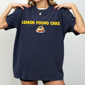 Puede incluir: Camiseta azul marino con la frase "LEMON POUND CAKE" en letras amarillas. Debajo, una ilustración de una rebanada de bizcocho con arándanos y una pequeña bandera. La camiseta tiene mangas cortas y cuello redondo.