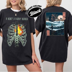 Op de afbeelding: Zwart Comfort Colors T-shirt met een afbeelding van een ribbenkast met een vlammend hart en de tekst "A Heart's A Heavy Burden" op de voorkant. De achterkant toont een Japanse landschap met een kasteel en een figuur.