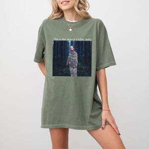 May include: Olive green t-shirt with a graphic featuring a man in camouflage in a forest setting. The text "This is the skin of a killer, bella" is above the graphic. The shirt has short sleeves and a relaxed fit.