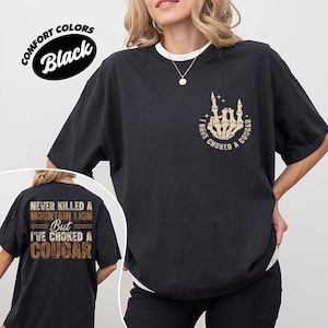 May include: Black t-shirt with the text "I'VE CHOKED A COUGAR" and a skeleton hand graphic. The back of the shirt reads "NEVER KILLED A MOUNTAIN LION BUT I'VE CHOKED A COUGAR". The shirt is a comfort colors brand.
