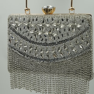 May include: Silver beaded clutch with a gold handle and a large crystal clasp. The rectangular bag is covered in small silver beads and larger crystal accents, with a fringe of silver beads hanging from the bottom.