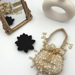 May include: A gold-toned, ornate framed mirror reflects a small beaded bag and a black velvet flower. A cream-colored, ring-shaped vase and a beaded bag with pearl accents are also present. The bag is embellished with sequins and pearls.