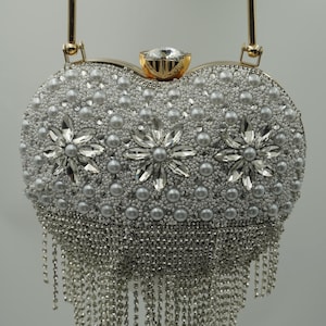 May include: A heart-shaped, silver beaded clutch with a gold handle. The clutch is embellished with pearls, rhinestones, and dangling silver chains. The clasp is a large, clear rhinestone set in gold.