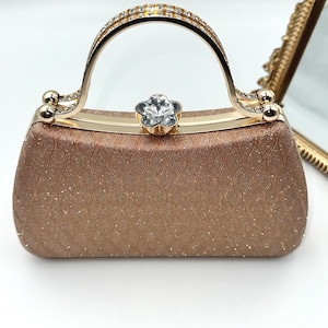 May include: A rectangular, glittery bronze clutch with a gold handle and clasp. The handle is adorned with rhinestones. A flower-shaped crystal embellishes the clasp. The clutch has a textured pattern and is likely for formal events.