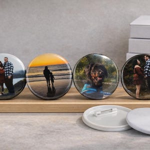 May include: Four round, glossy photo buttons with images of a couple, a sunset, and a dog, displayed on a wooden board. Two blank white buttons and a stack of white boxes are also visible.