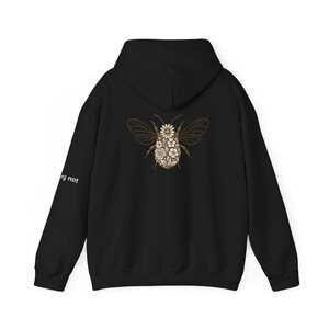 Puede incluir: Sudadera con capucha negra con un diseño floral de abeja en la espalda. El cuerpo de la abeja está lleno de flores blancas y crema. La sudadera también tiene el texto "worry not" impreso en la manga izquierda.