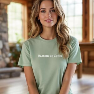 May include: A pale green t-shirt with the white text "Bean me up Coffee". The shirt has short sleeves and is made of a soft material. The person is sitting in a room with wooden features.