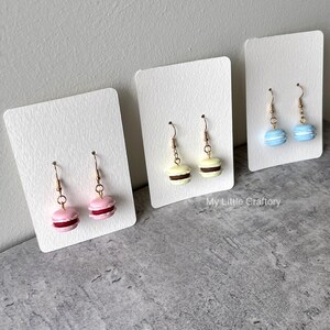 Könnte beinhalten: Drei Paar Macaron-Ohrringe auf weißen Präsentationskarten. Die Ohrringe haben goldfarbene Haken und Miniatur-Macarons in Rosa, Gelb und Blau. Die rosa Macarons haben eine dunkelrote Füllung, die gelben eine braune Füllung und die blauen eine weiße Füllung.