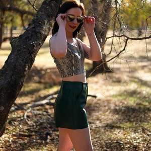 May include: A woman wearing a snakeskin print crop top and a dark green leather skirt. She is standing in a wooded area and is holding sunglasses in her hands.