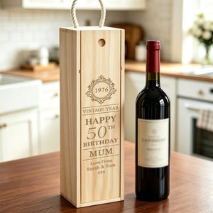 Könnte beinhalten: Eine helle Weinkiste aus Holz mit einem Seilgriff und einer Gravur mit der Aufschrift 1976, VINTAGE YEAR, HAPPY 50TH BIRTHDAY MUM, Love from Sarah & Tom, XXX. Eine dunkle Rotweinflasche steht neben der Kiste auf einer Holzoberfläche.