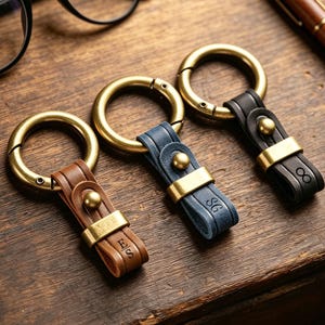 May include: Three leather keychains in brown, blue, and black, each featuring a brass circular ring, a brass stud, and a brass loop. The brown keychain is embossed with ES, the blue with SG, and the black with an infinity symbol.
