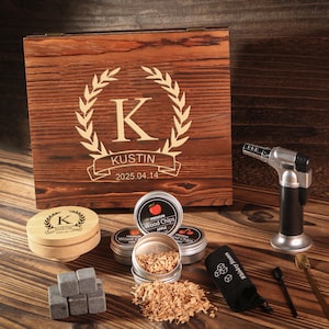 May include: A wooden box with a laser-engraved design, including a large letter "K" within a laurel wreath, the name "Kustin", and the date "2025.04.14". The box is surrounded by smoking accessories, including wood chips, a torch, and whiskey stones.