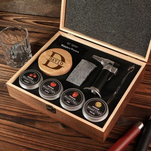 May include: A wooden gift box set with whiskey accessories. Includes a personalized wooden coaster, four tins of wood chips, whiskey stones, a torch lighter, and a mixing spoon. A rocks glass sits to the left.