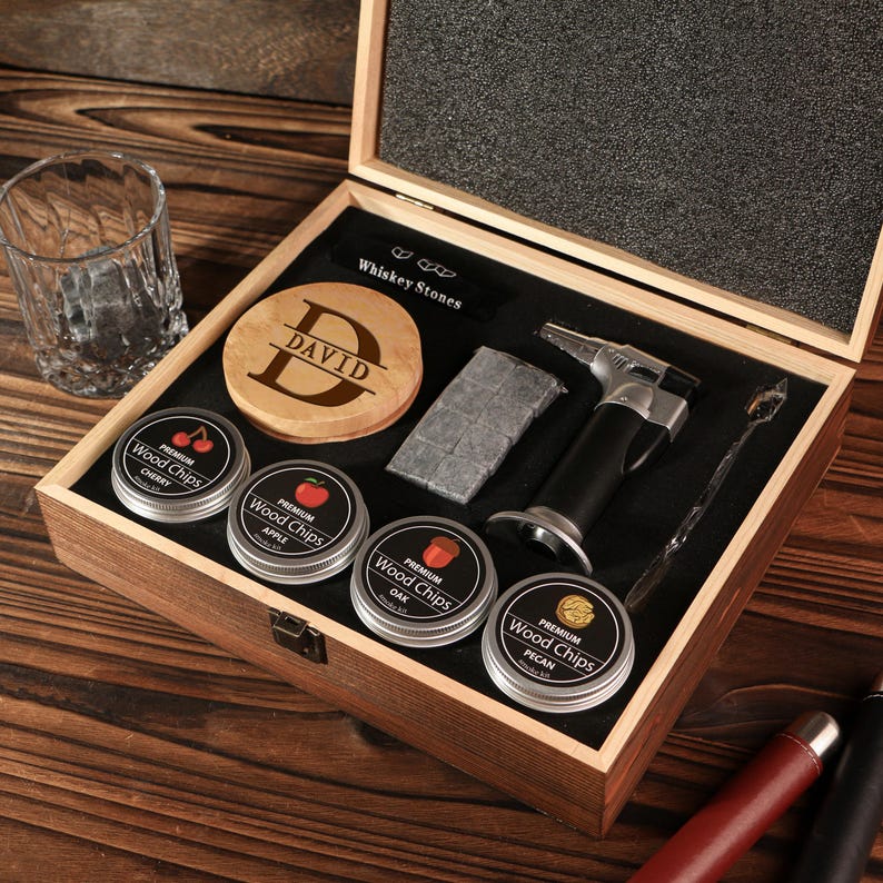 May include: A wooden gift box set with whiskey accessories. Includes a personalized wooden coaster, four tins of wood chips, whiskey stones, a torch lighter, and a mixing spoon. A rocks glass sits to the left.