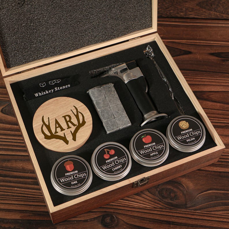 May include: A wooden gift set for whiskey enthusiasts. The set includes whiskey stones, a torch, a stirring spoon, a wooden coaster with "HARY" and antlers, and tins of wood chips labeled "Oak", "Cherry", "Apple", and "Pecan".