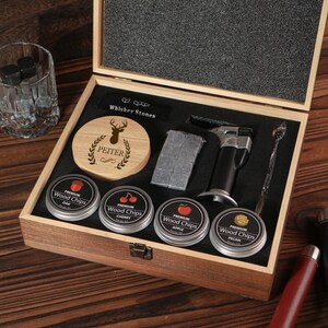 May include: A wooden gift set for whiskey enthusiasts, featuring a wooden box with various accessories. Includes whiskey stones, a torch, a stirring spoon, and tins of wood chips. The box also contains a coaster with a deer design and the name "PEITER".