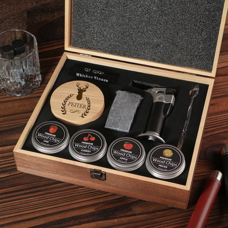 May include: A wooden gift set for whiskey enthusiasts, featuring a wooden box with various accessories. Includes whiskey stones, a torch, a stirring spoon, and tins of wood chips. The box also contains a coaster with a deer design and the name "PEITER".