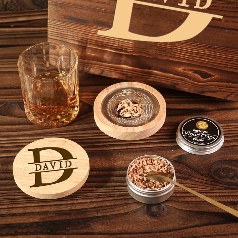 May include: A wooden coaster with the name "DAVID" in a bold font, a rocks glass with amber liquid, a wood smoker with wood chips, and a tin of pecan wood chips. The items are arranged on a wooden surface.
