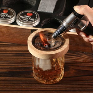 May include: A wooden cocktail smoker kit with a glass and a torch being used to light wood chips. The kit includes two tins of wood chips, one labeled "Oak" and the other "Apple". The glass has a decorative pattern.