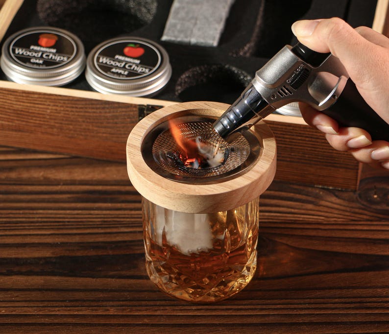 May include: A wooden cocktail smoker kit with a glass and a torch being used to light wood chips. The kit includes two tins of wood chips, one labeled "Oak" and the other "Apple". The glass has a decorative pattern.