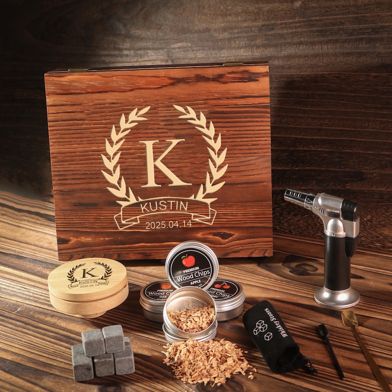May include: A wooden box with a laser-engraved design, including a large letter "K" within a laurel wreath, the name "Kustin", and the date "2025.04.14". The box is surrounded by smoking accessories, including wood chips, a torch, and whiskey stones.