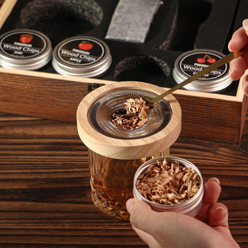 May include: A wooden cocktail smoker kit with a glass, wood chips, and a metal spoon. The kit includes small tins labeled "Wood Chips" with flavors like oak, apple, and cherry. The smoker has a wooden top and a metal mesh filter.