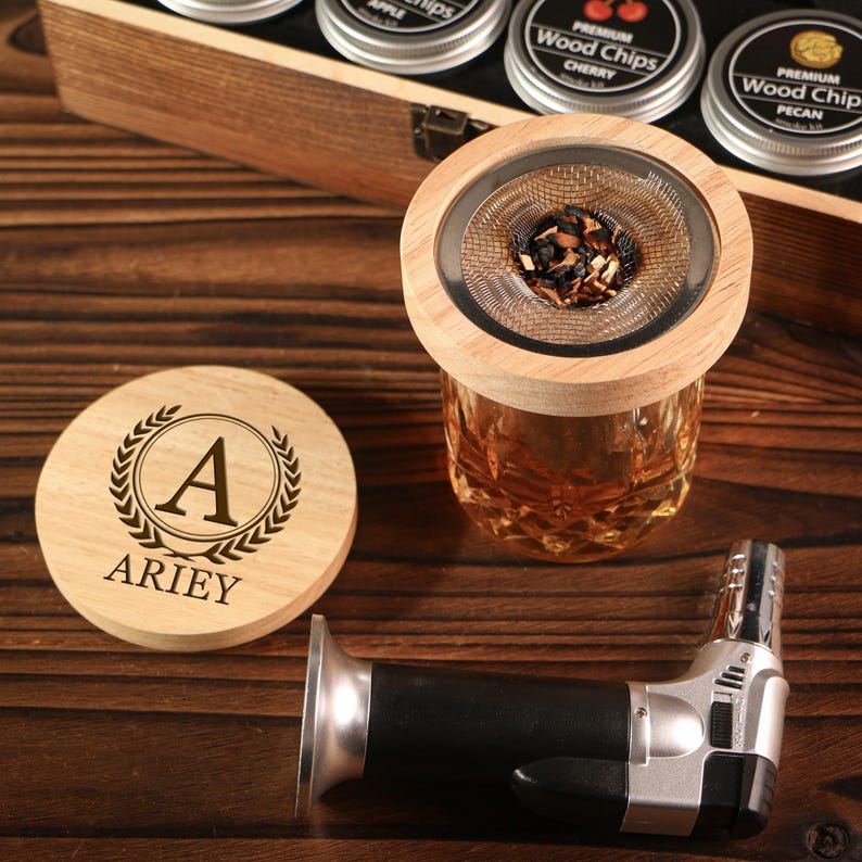 May include: A wooden coaster with the name "ARIEY" and the letter "A" inside a laurel wreath, a glass with a wooden lid and a metal mesh, a black and silver lighter, and a wooden box with small metal containers.