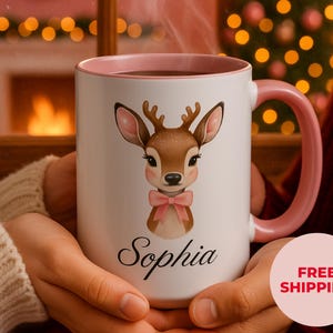May include: White ceramic mug with a pink interior and handle. The mug features a watercolor illustration of a deer with a pink bow and the name "Sophia" in script. The mug is held by a person.