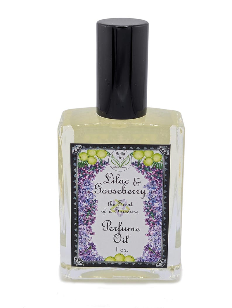 Op de afbeelding: Een heldere glazen parfumolie fles met een zwarte dop. Het etiket luidt "Lilac & Gooseberry, the Scent of a Sorceress, Perfume Oil, 1 oz." met bloemen- en fruitillustraties. De fles bevat een lichtgele vloeistof.