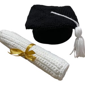 May include: A crocheted graduation set comprising a black mortarboard with a white tassel and a rolled-up white diploma tied with a gold ribbon. Ideal for celebratory events.