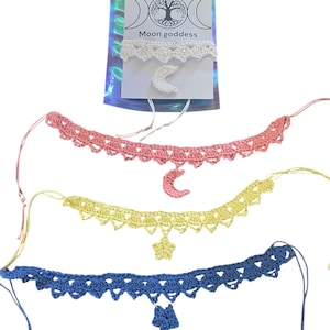 Pode incluir: Uma coleção de gargantilhas de crochê em rosa, amarelo e azul, cada uma com um charme único. A rosa apresenta uma lua crescente, a amarela uma estrela e a azul uma estrela. Uma gargantilha branca com um pingente de lua crescente também é visível, com o texto "Moon goddess".