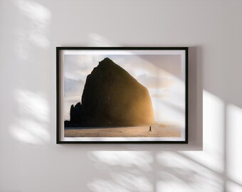 May include: A framed landscape photograph featuring a large, dark, monolithic rock formation on a sandy beach at sunset. The golden light illuminates the side of the rock, while a small silhouette of a person walks along the shoreline below.
