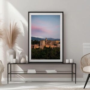 K&ouml;nnte beinhalten: Gerahmter Druck des Alhambra-Palastes in Granada, Spanien, bei Sonnenuntergang. Das Bild zeigt die historische Architektur, die vor einem rosa-blauen Himmel beleuchtet wird, mit schneebedeckten Bergen im Hintergrund. Der Druck befindet sich auf einem schwarzen Metalltisch.
