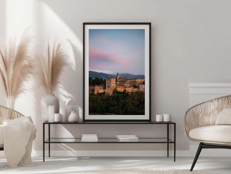 K&ouml;nnte beinhalten: Gerahmter Druck des Alhambra-Palastes in Granada, Spanien, bei Sonnenuntergang. Das Bild zeigt die historische Architektur, die vor einem rosa-blauen Himmel beleuchtet wird, mit schneebedeckten Bergen im Hintergrund. Der Druck befindet sich auf einem schwarzen Metalltisch.