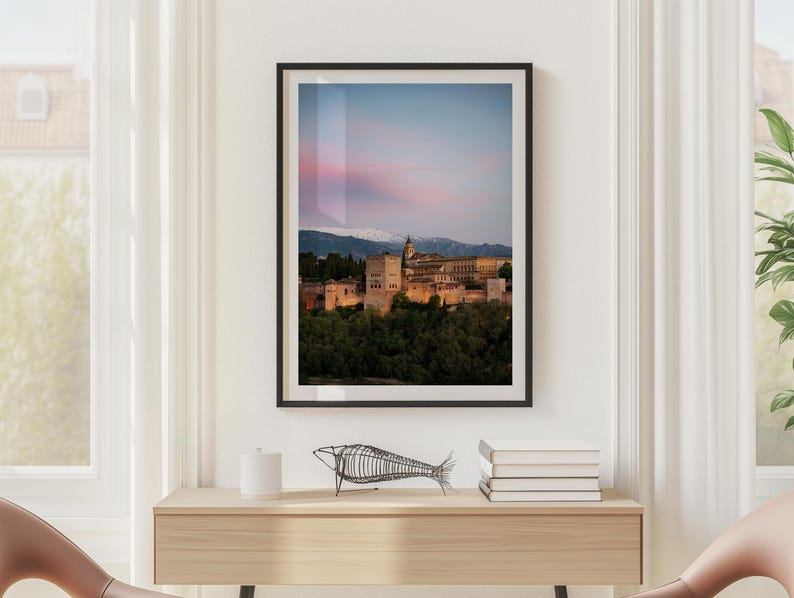 K&ouml;nnte beinhalten: Gerahmter Druck des Alhambra-Palastes in Granada, Spanien, in der Abendd&auml;mmerung. Das Bild zeigt den beleuchteten Palast vor dem Hintergrund eines rosa-blauen Himmels und schneebedeckter Berge. Der Druck wird auf einer hellen Holzkonsolentisch pr&auml;sentiert.