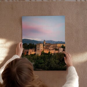 K&ouml;nnte beinhalten: Quadratischer Leinwanddruck des Alhambra-Palastes in Spanien, gehalten von einer Person. Das Bild zeigt den beleuchteten Palast in der Abendd&auml;mmerung, mit einem rosa-blauen Himmel und schneebedeckten Bergen im Hintergrund. Der Druck wird auf einer beigen, gewebten Oberfl&auml;che pr&auml;sentiert.