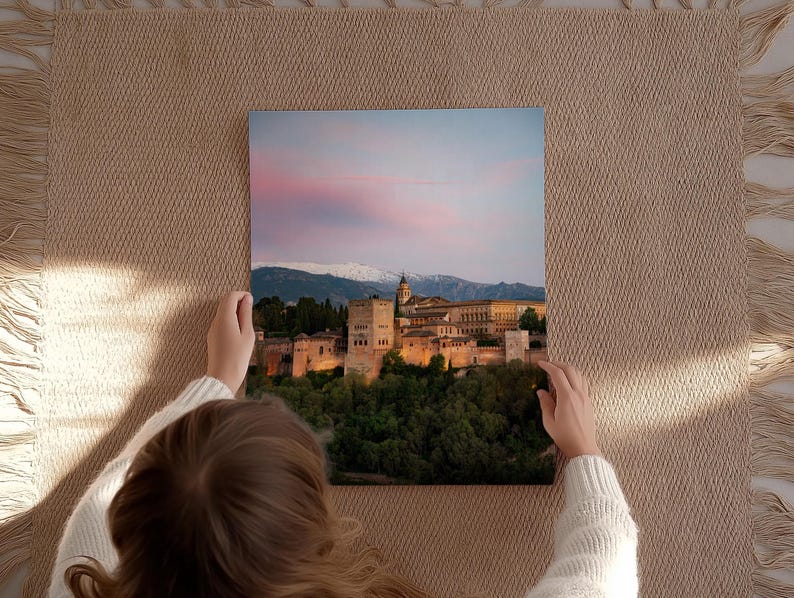 K&ouml;nnte beinhalten: Quadratischer Leinwanddruck des Alhambra-Palastes in Spanien, gehalten von einer Person. Das Bild zeigt den beleuchteten Palast in der Abendd&auml;mmerung, mit einem rosa-blauen Himmel und schneebedeckten Bergen im Hintergrund. Der Druck wird auf einer beigen, gewebten Oberfl&auml;che pr&auml;sentiert.