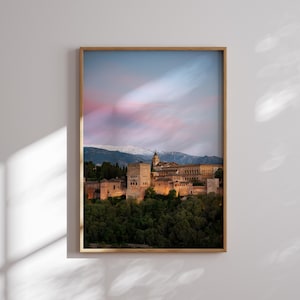 K&ouml;nnte beinhalten: Gerahmtes Foto eines historischen Burgkomplexes in der Abendd&auml;mmerung. Das Bild zeigt eine warme Farbpalette mit dem beleuchteten Schloss vor dem Hintergrund eines rosa-blauen Himmels und schneebedeckter Berge. Der Rahmen ist hellbraun.