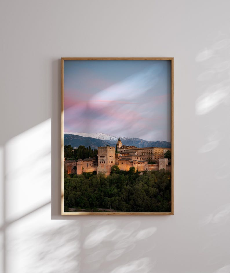 K&ouml;nnte beinhalten: Gerahmtes Foto eines historischen Burgkomplexes in der Abendd&auml;mmerung. Das Bild zeigt eine warme Farbpalette mit dem beleuchteten Schloss vor dem Hintergrund eines rosa-blauen Himmels und schneebedeckter Berge. Der Rahmen ist hellbraun.