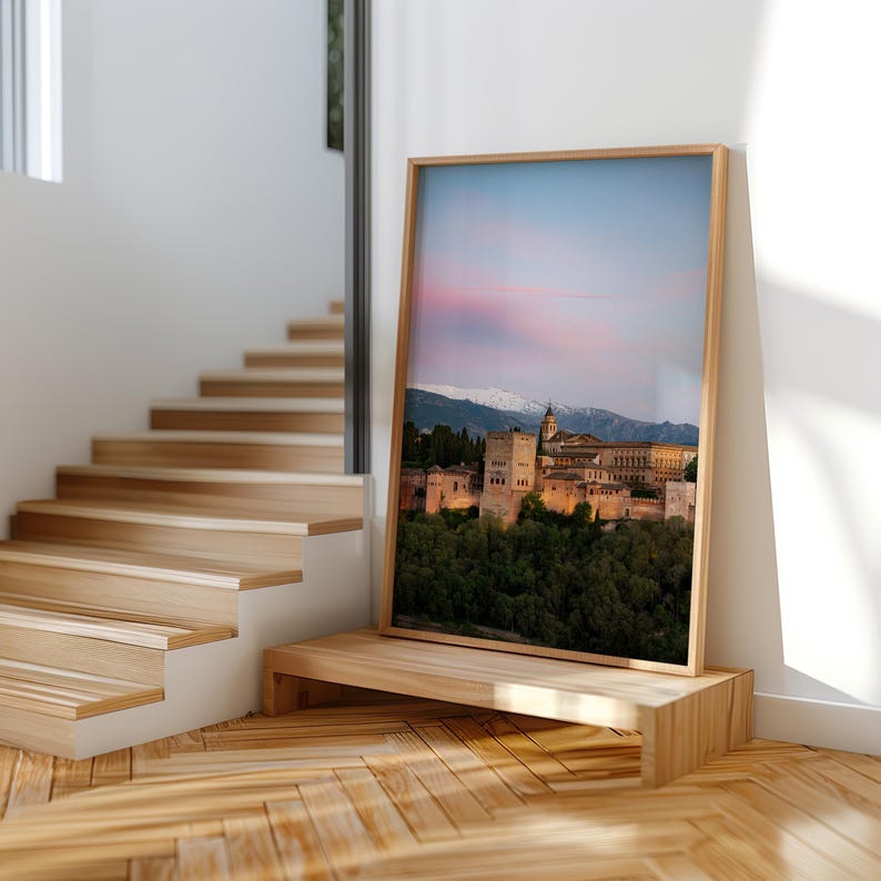 K&ouml;nnte beinhalten: Ein gerahmter Druck des Alhambra-Palastes in Granada, Spanien, bei Sonnenuntergang. Das Bild zeigt die historische Architektur, gr&uuml;ne B&auml;ume und schneebedeckte Berge unter einem rosa und blauen Himmel. Der Rahmen ist aus hellem Holz.