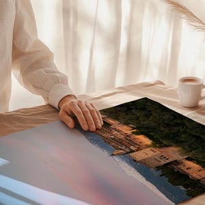 K&ouml;nnte beinhalten: Ein Landschaftsdruck mit einem Geb&auml;ude vor einem Bergpanorama, der auf einem Tisch ausgestellt ist. Der Druck ist rechteckig und hat einen farbenfrohen Himmel. Eine Hand ruht auf dem Druck. Eine wei&szlig;e Tasse mit Kaffee und eine Vase mit dekorativem Gras stehen ebenfalls auf dem Tisch.