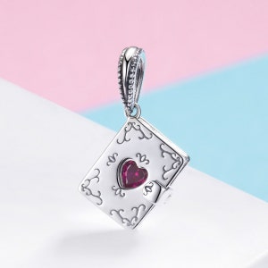 May include: A silver charm shaped like a closed book with a red heart in the centre. The book has an ornate design and a clasp.