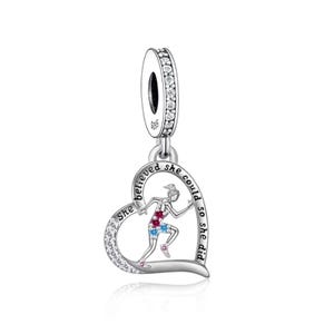 Könnte beinhalten: Ein silberner Charm-Anhänger mit herzförmigem Anhänger, der eine laufende Figur zeigt. Das Herz ist mit klaren Steinen umrandet und trägt die Worte „She believed she could so she did“. Der Charm hat eine mit klaren Steinen besetzte Schlaufe.