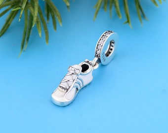 May include: A silver charm in the shape of a white and light blue trainer with a lace-up design. The charm has a silver loop with clear stones. The background is a solid blue.