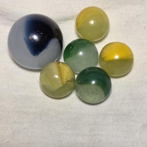 May include: A collection of colorful marbles. One large marble is gray with a dark blue swirl. The other marbles are yellow, green, and yellow-green, with various patterns and swirls. The marbles are arranged on a white surface.